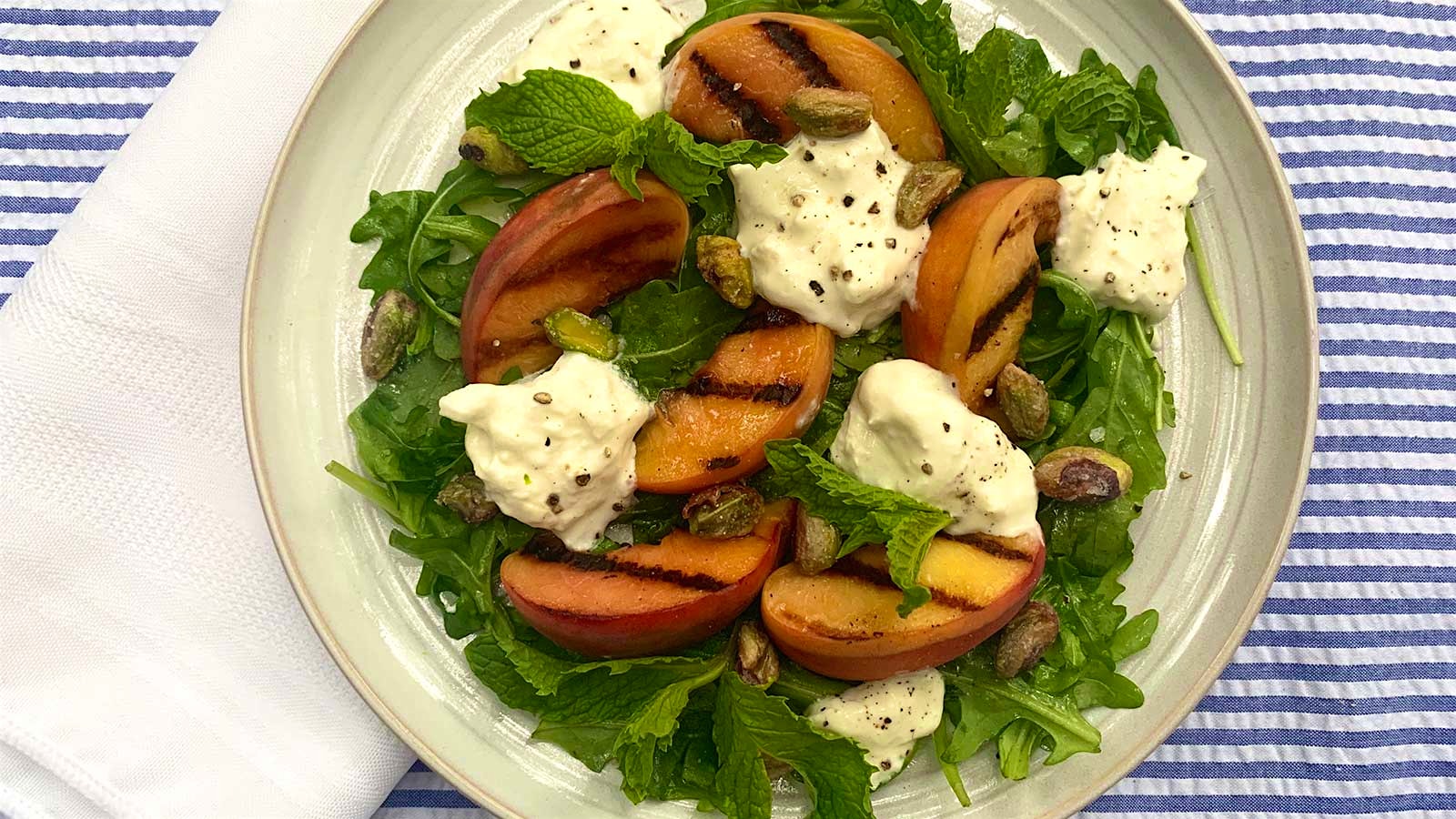  Plate of salad with grilled peach slices and dollops of burrata cheese on top of lettuce, garnished with pistachios and mint